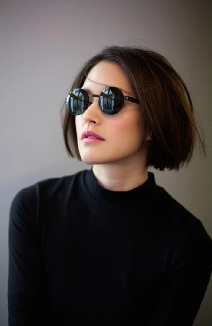 hairstyles for short hair, woman with round black glasses, and black turtleneck, with straight brown bob, parted at the side