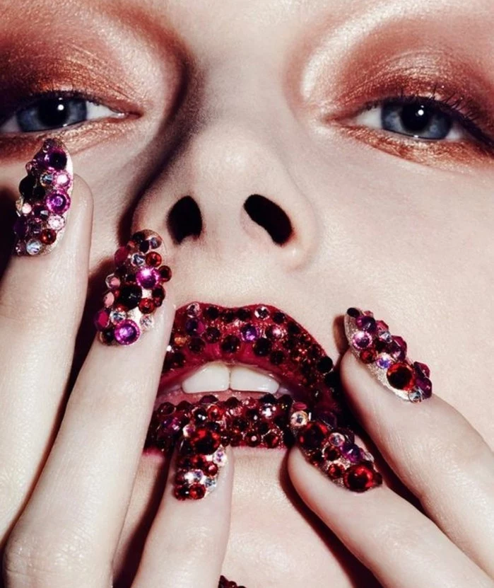 extreme close up of woman's face and fingers, brown eye make up, red lips covered with pink and purple rhinestones, fingernails covered with rhinestones in the same color