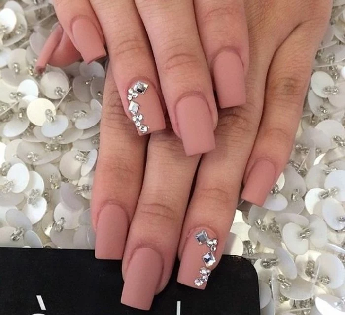 two hands with square nails, painted with dark nude matte polish, two of them decorated with round and square rhinestones