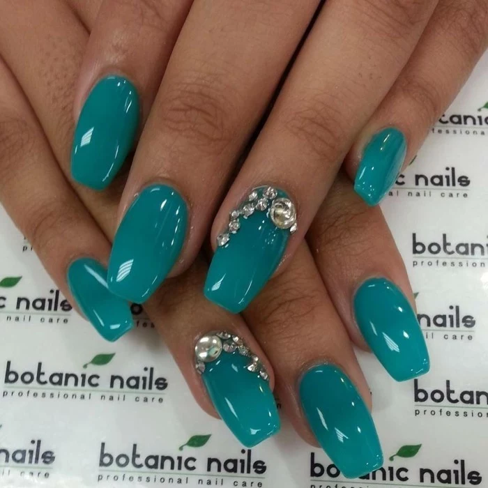 close up of tan hand with semi-square nails, painted in deep turquoise, two of the nails are decorated with silver rhinestones
