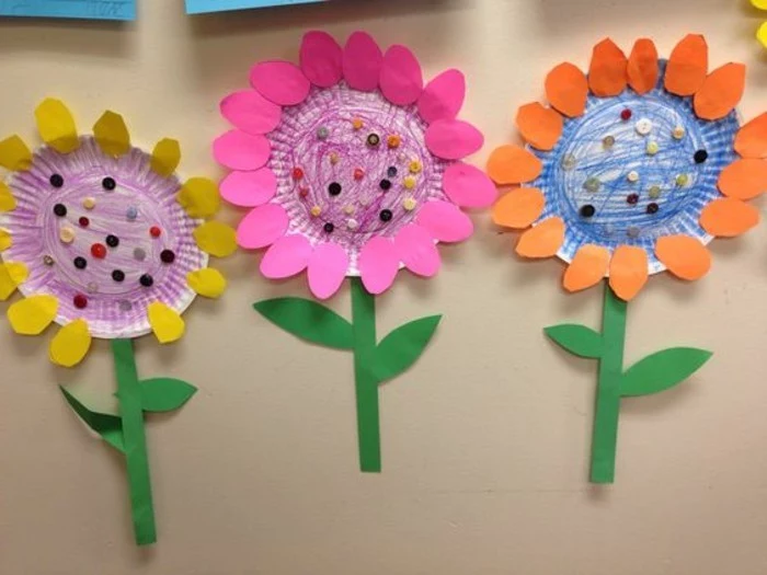 craft ideas for kids, three flowers made from paper plates, colored with pencils, with stalks made from green paper and petals made from yellow, pink and orange paper
