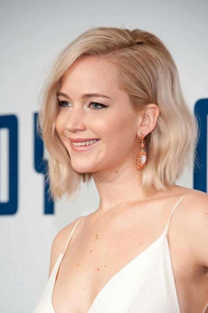 close up of jennifer lawrence, with wavy light blonde side parted bob, wearing white strappy top and orange earrings
