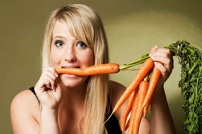 dark blue eyes, blonde woman with long hair and side bangs, holding a bunch of carrots in one hand, and biting onto one