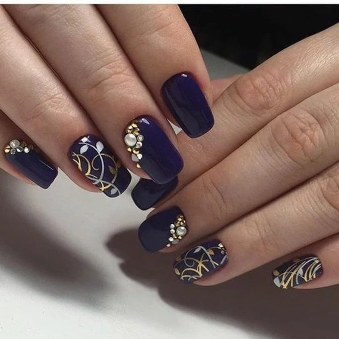 two hands with violet nail polish, several nails decorated with gold and silver drawings, rhinestones and pearls