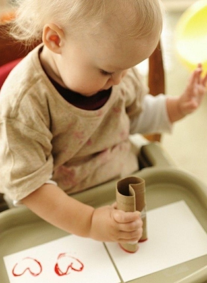 blond toddler in grey shirt, using a paper roll to stamp red heart-shapes on two white pieces of paper, placed on a plastic tray