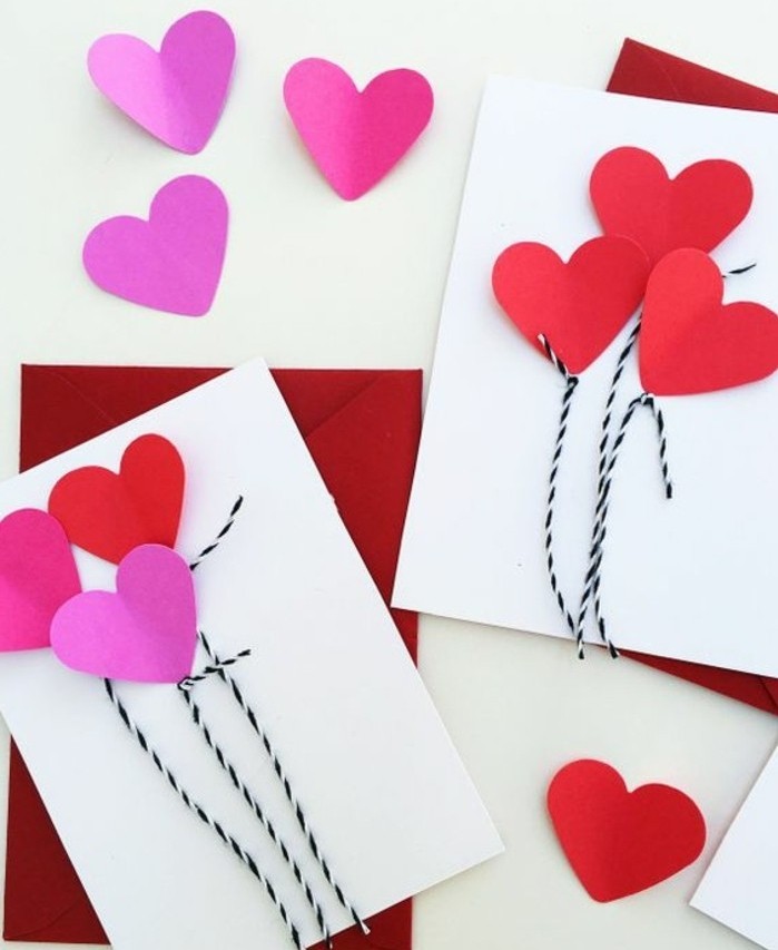 two white cards, decorated with pink and red heart shapes, and black and white thread, placed over two red envelopes, more pink and red heart shapes nearby