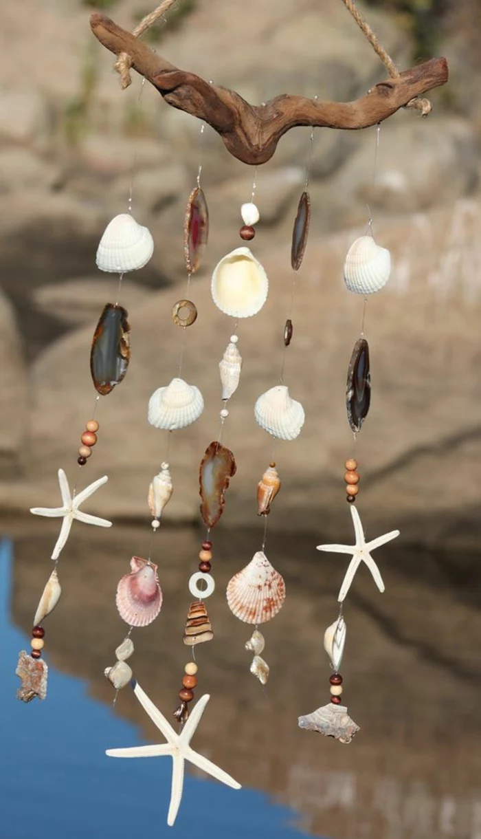 summer diys, wind chime made from multicolored seashells, pebbles and starfish, tied together with clear plastic string, hanging from a piece of driftwood