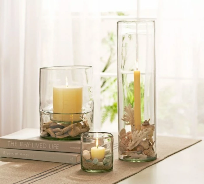 summer craft ideas, three glass containers in different shapes and sizes, containing either pebbles, small pieces of driftwood or corals, and a lit candle