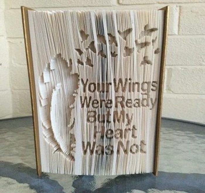 folded book art, thick closed book, with light brown hard covers, and a long message carved into its pages, along with a feather and some small bird shapes