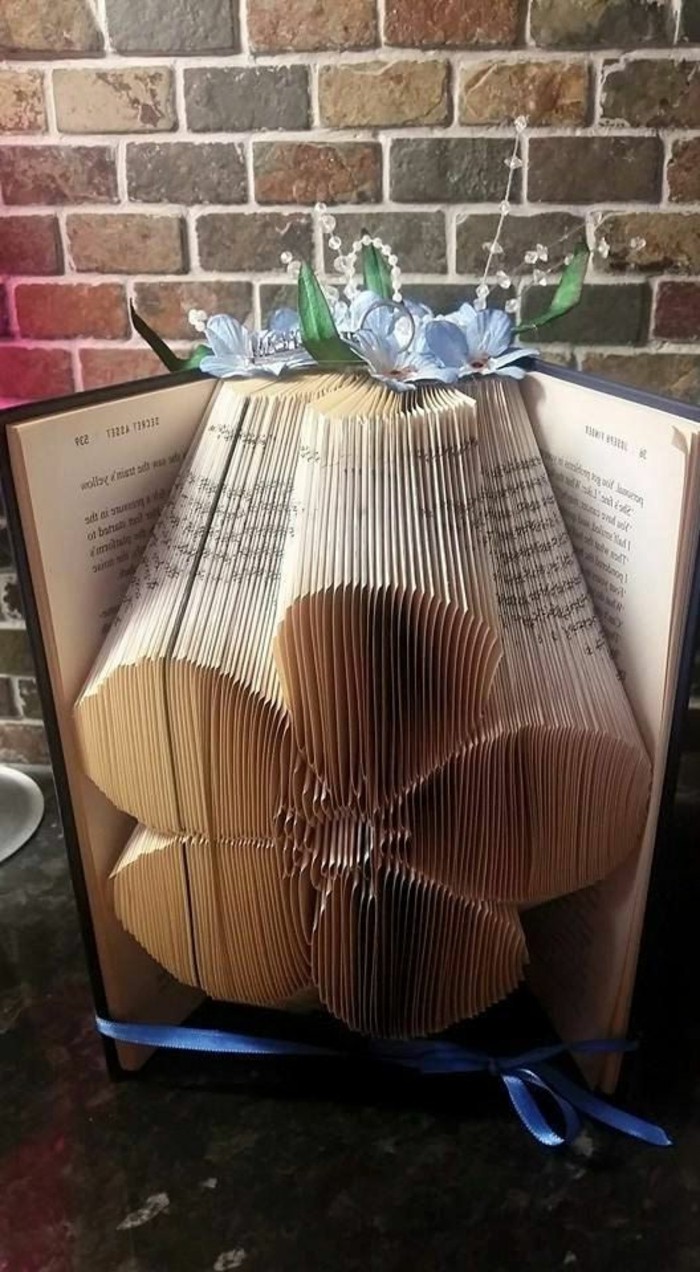 folded book art, flower made from folded pages, inside an open book, with hard black covers, tied with blue ribbon, and decorated with blue paper flowers