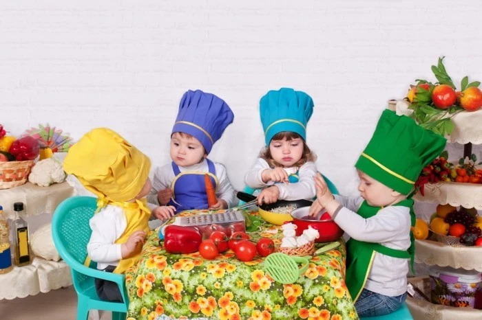 four toddlers wearing chef's hats and aprons in different colors, sitting on small plastic chairs around a table, with various cooking utensils and vegetables