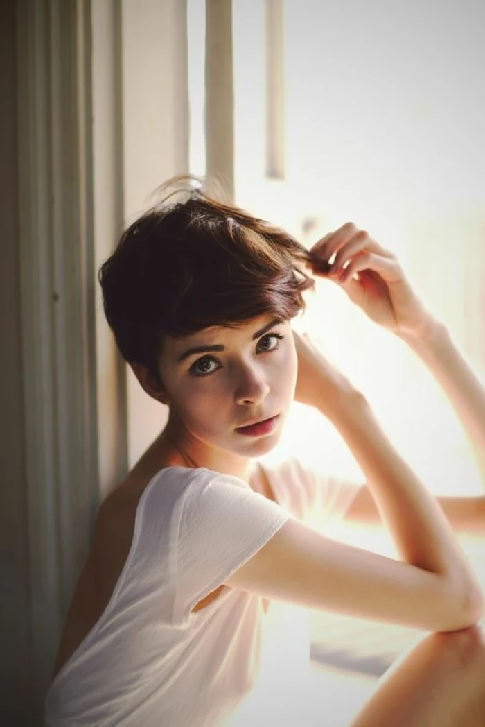 pixie cut, slim girl with big eyes, and very short wavy brown hair, longer bangs swept to one side on her forehead, wearing white open-back t-shirt