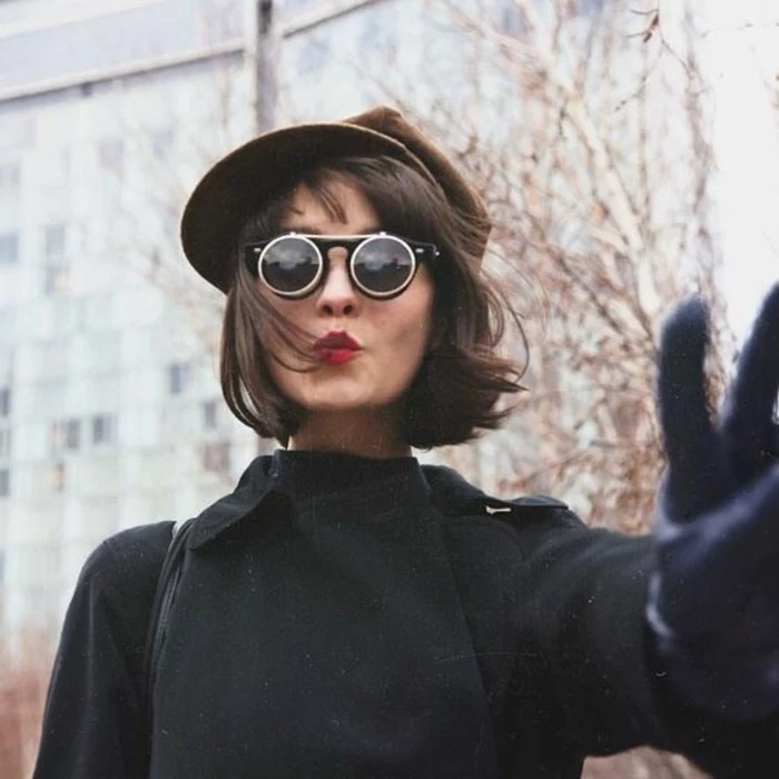 woman with wind-swept, chin-length brown hair and dark round sunglasses, wearing brown cap and black coat, making the peace sign, with one gloved hand
