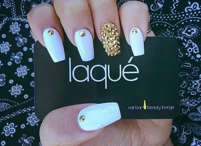 hand with long square nails, four painted white and decorated with a single golden rhinestone, one entirely covered in gold rhinestones, holding black business card