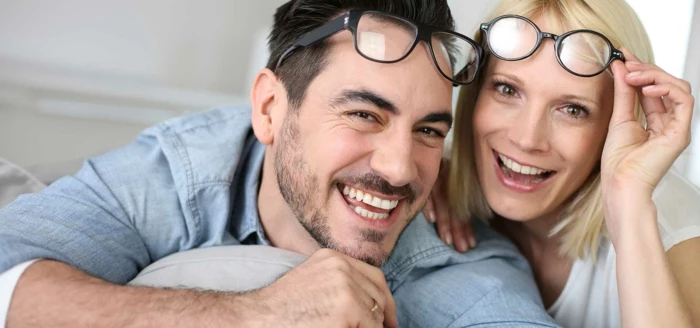 hazel eye color, laughing man with dark hair, short beard and mustache, wearing pale denim shirt, with glasses over his head, next to a laughing blonde woman with white top, also with glasses on her head