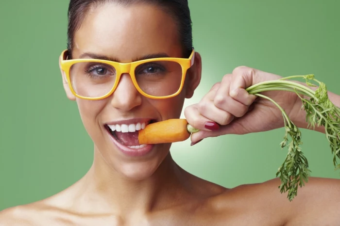 hazel eye color, smiling woman with big white teeth, brown hair tied back, wearing big glasses with yellow frames, holding and biting onto a carrot