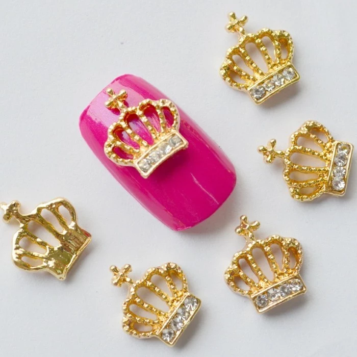 bling nail designs, hot pink fake nail, decorated with golden crown with rhinestones, next to several more crown decorations 