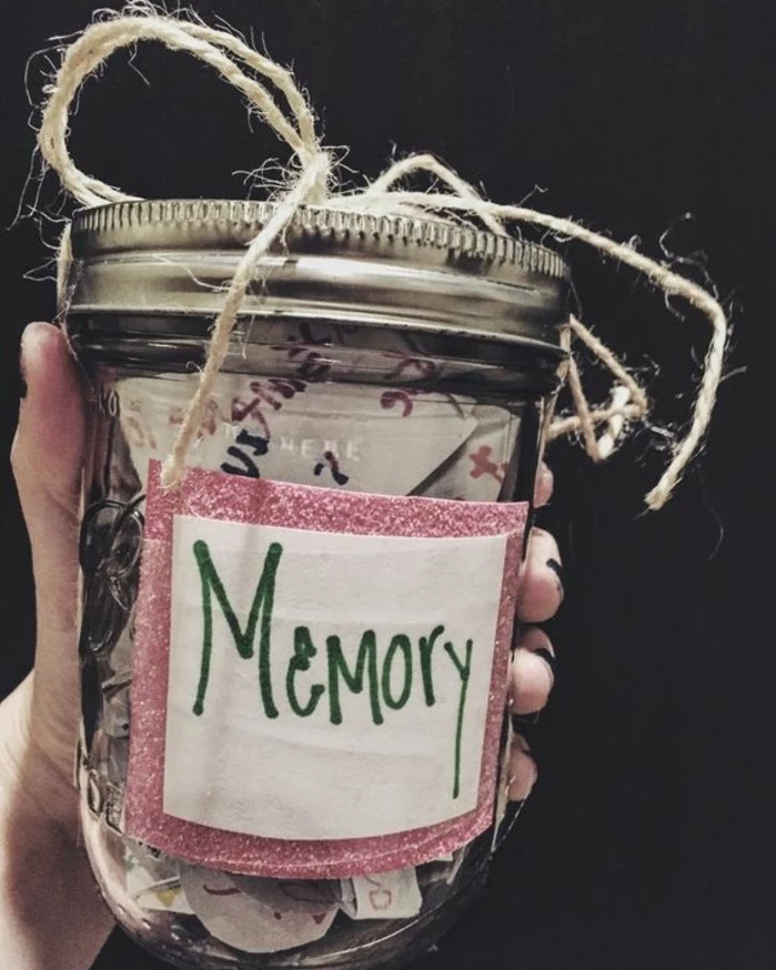 hand holding a clear mason jar, with silver lid, filled with colorful bits of paper, tied with simple string, with a white and pink label saying memory in green