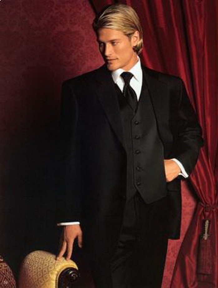 blonde man with hair tucked behind his ears, wearing a formal black suit with vest and tie