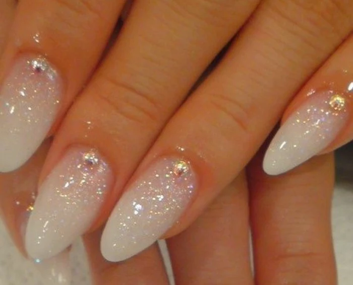 close up of four sharp nails, painted in white polish, and covered in light glitter, with one rhinestone each