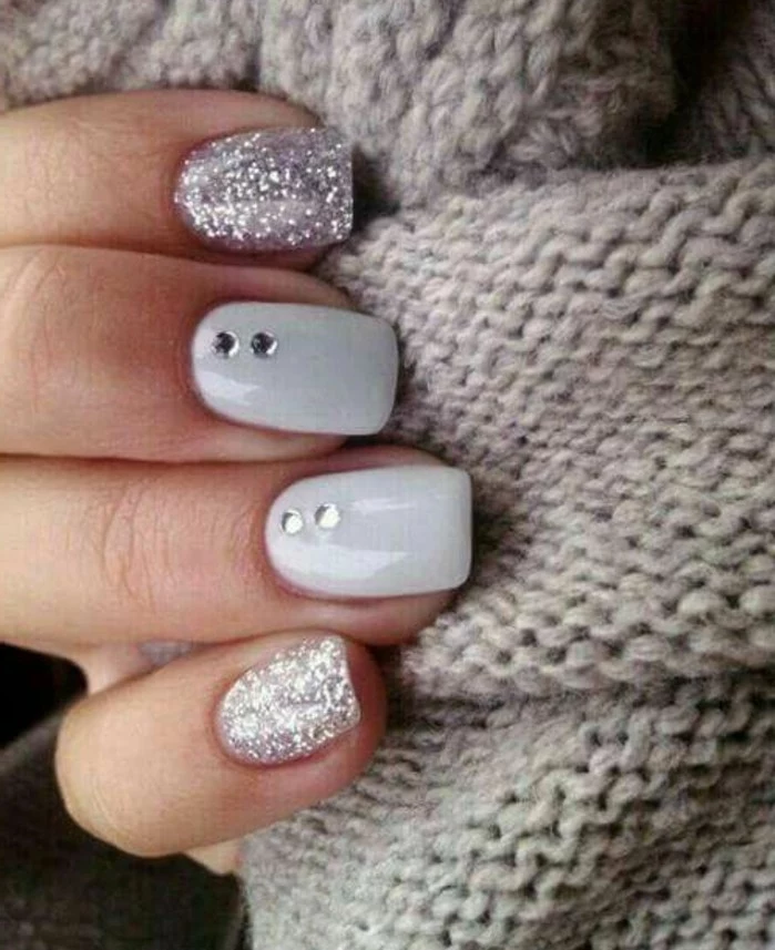 nail designs with rhinestones and glitter, close up of four fingers on grey woolen fabric, two nails have silver glitter polish, two are grey with rhinestone decoration