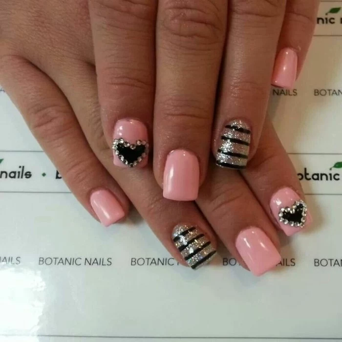hand with square nails painted in pink, two nails decorated with black hearts and rhinestones, other two nails decorated with gold glitter and black stripes