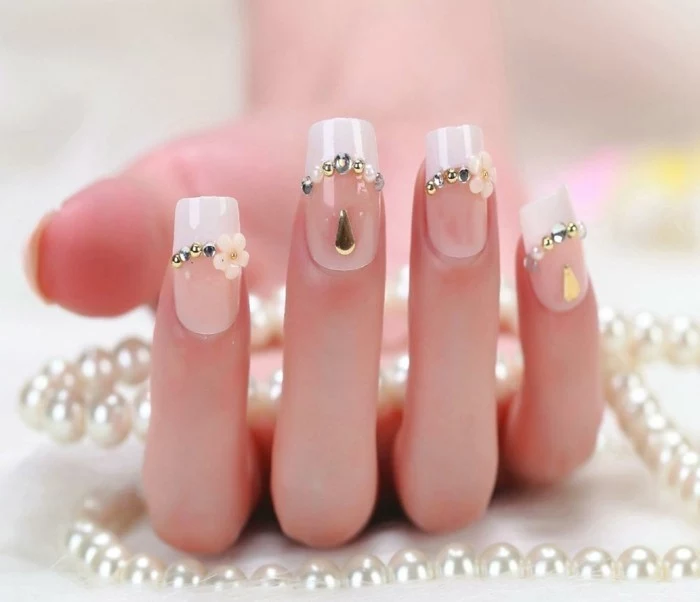 nail designs with rhinestones, hand with french manicure, decorated with silver and white rhinestones, golden teardrop-shaped stickers and acrylic flowers, holding pearl necklace