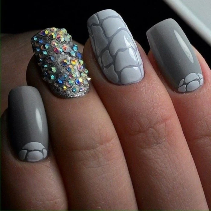 nail designs with rhinestones, close up of four nails, three dark grey with light grey details, one sparkling grey and decorated with many multicolored rhinestones