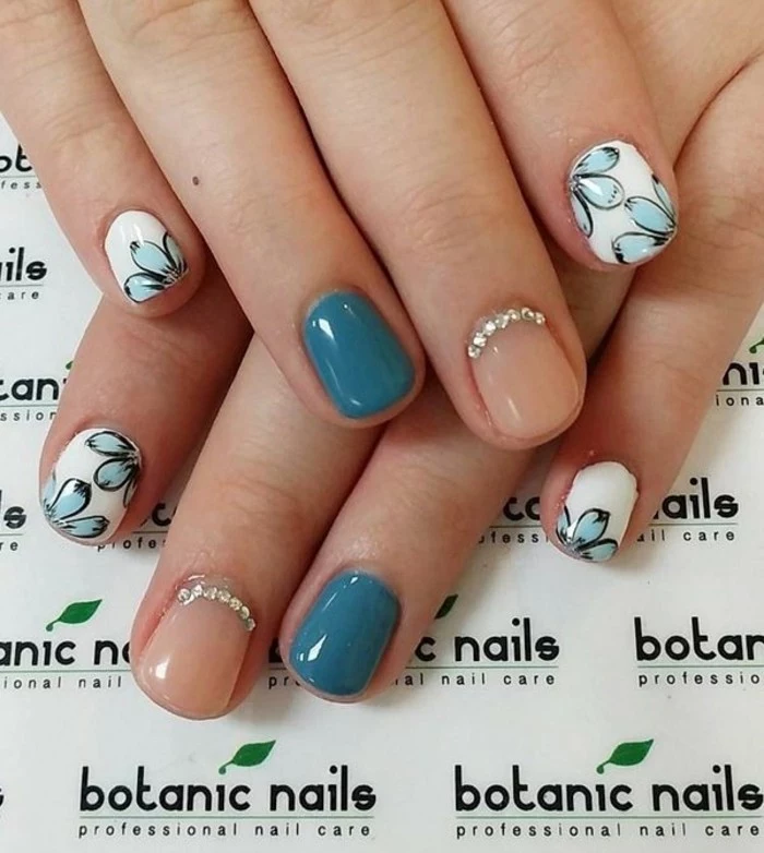 nails with rhinestones, two hands, one resting on top of another, nails in pastel pink with rhinestones and blue, pinkie and index finger nails white with pale blue flowers