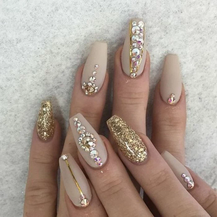 nude nails with rhinestones, close up of two hands with pale nude matte nail polish, two nails covered in gold glitter, the rest have rhinestones 
