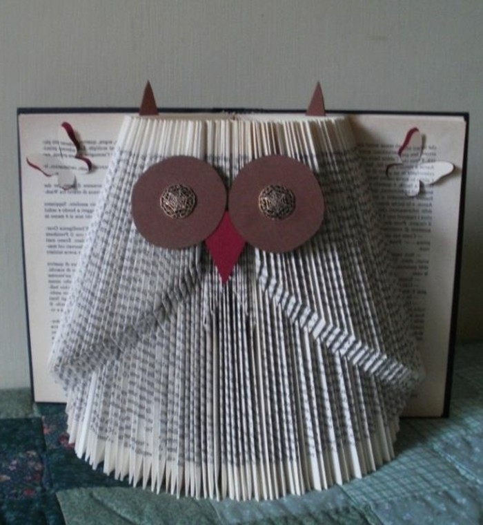 owl ornament decorated with felt cutouts and metal buttons, made from folded pages, inside an open book with dark hard covers, and butterfly-shaped cutouts