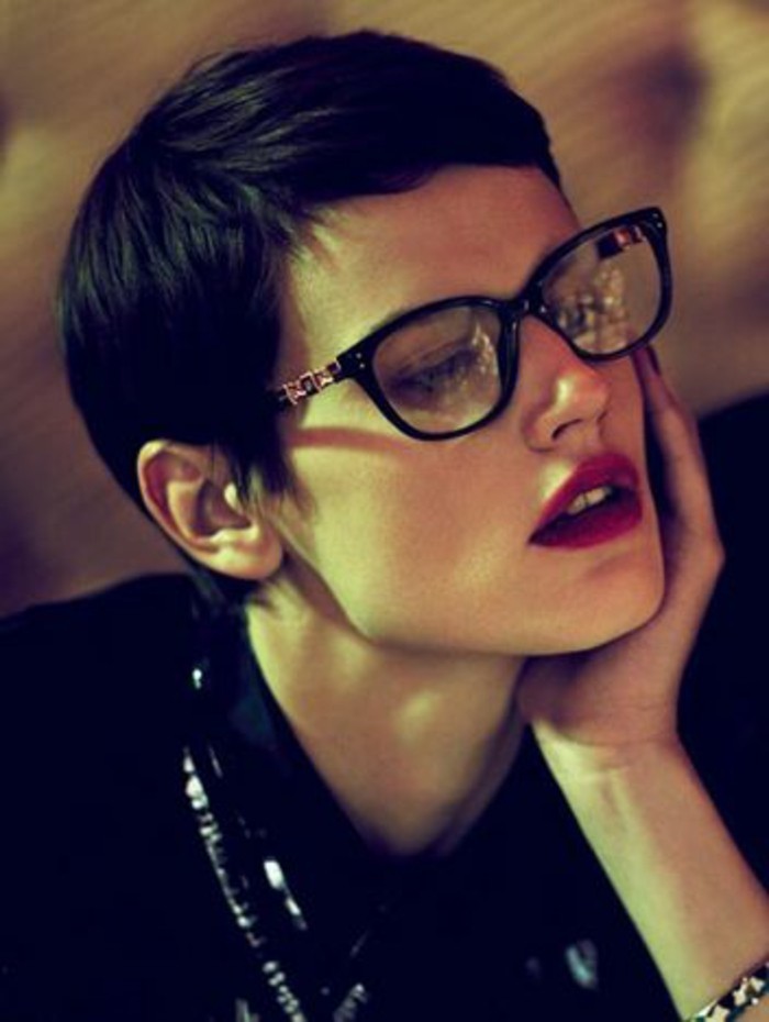 dark-haired woman, with very short pixie cut, wearing glasses with black frames, and bright red lipstick, leaning on one hand