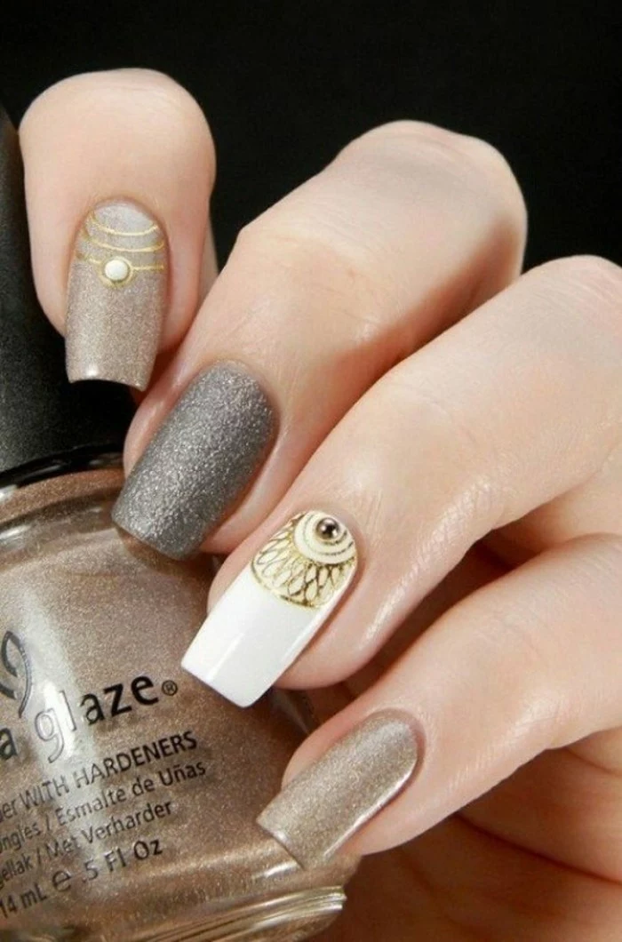 rhinestone nail art, hand holding bottle of pale brown shimmering nail polish, hand's nails painted in similar colors, with gold details, one rhinestone and a white accent