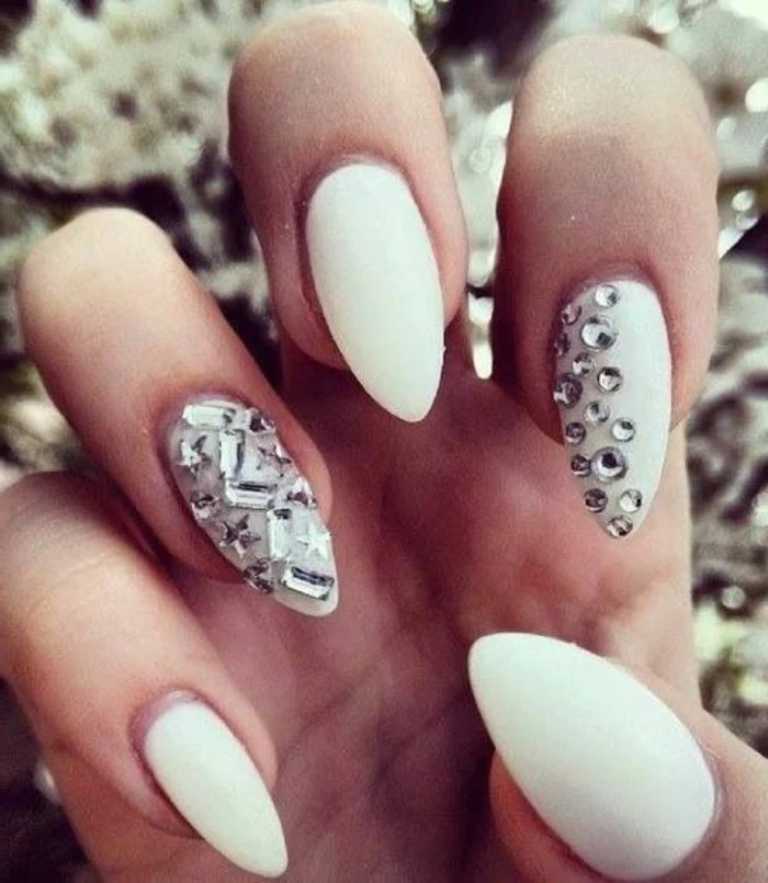 nail designs with rhinestones and glitter, close up of hands with sharp nails painted in milky white polish, two fingers decorated with round, star-shaped and rectangular rhinestones
