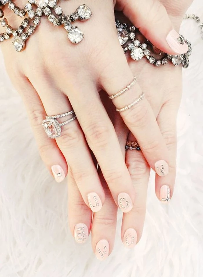nail designs with rhinestones and glitter, two hands with rings and bracelets, with round nails painted in pale pink polish, each decorated with several rhinestones