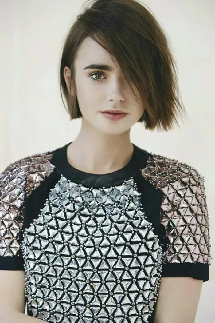 lily collins with short, dark brown bob, bangs covering her eye, wearing black top, with triangular silver ornaments