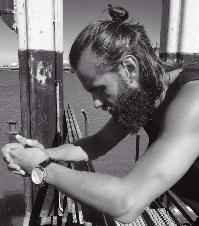 medium length hairstyles, bearded man leaning on railing, looking down and to the side, hair tied in man bun