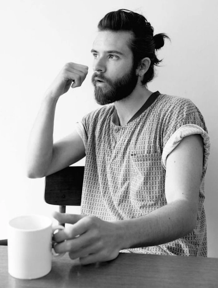 shoulder length hairstyles, bearded man sitting at a table, holding white mug, hair tied in man bun