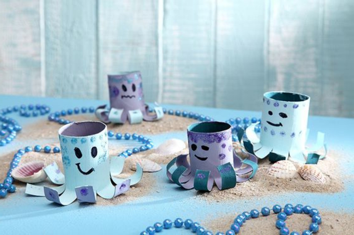 summer crafts, four octopi decorations, made from toilet paper rolls, painted in pale blue and violet, placed on blue surface, with seashells sand and blue beaded ropes