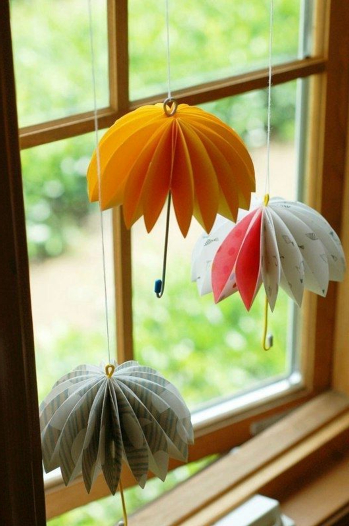summer craft ideas, three multicolored folded paper umbrella decorations, with wire handles, hanging from window pane, on white thread 