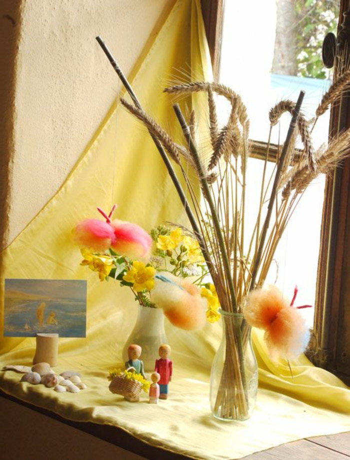 summer crafts, window settee decorated with bright yellow fabric, with two vases containing wheat stalks, yellow flowers and colorful woolen decorations, seashells and pebbles, a postcard and a small doll family nearby