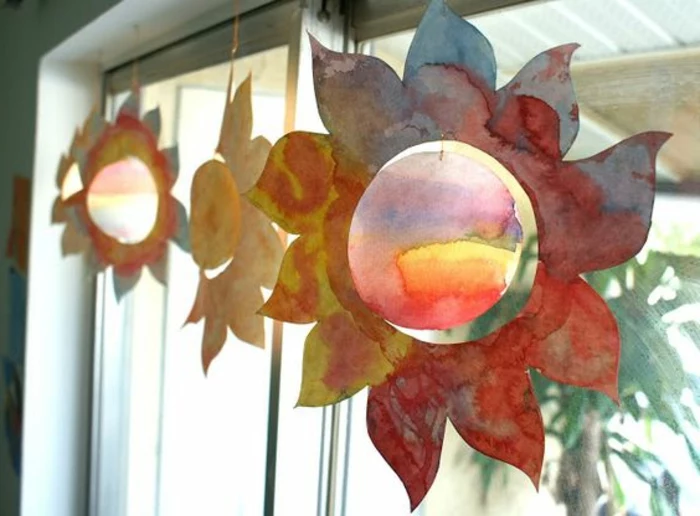 easy fun diys, several sun-shaped paper decorations, painted with different watercolors, hanging from a window