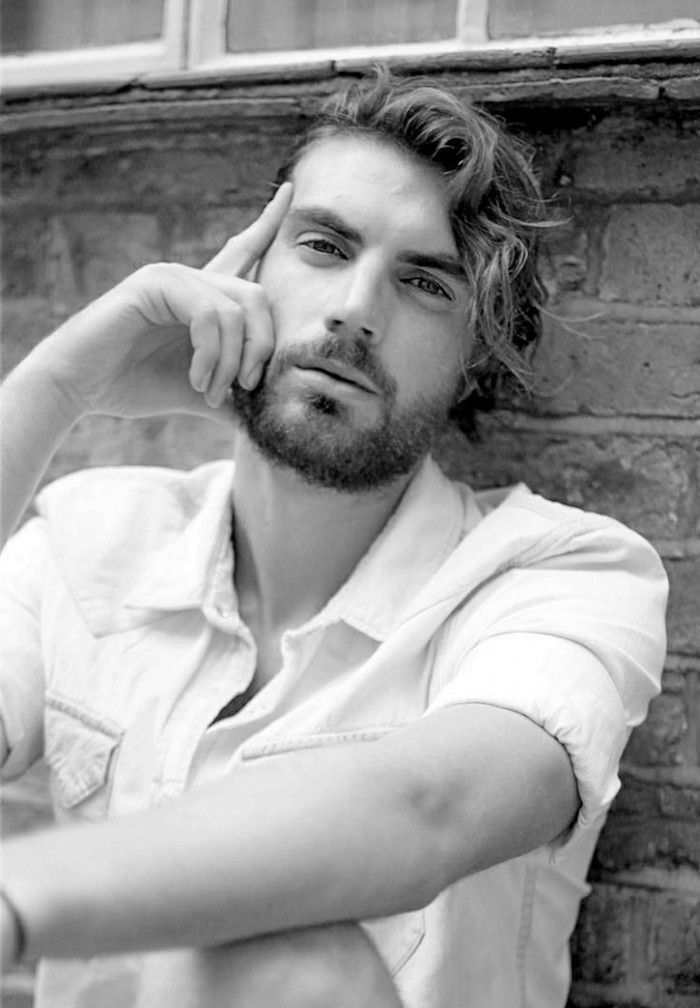 mid length hair, man leaning on brick wall, curly hair parted to one side, stubbly beard and white shirt