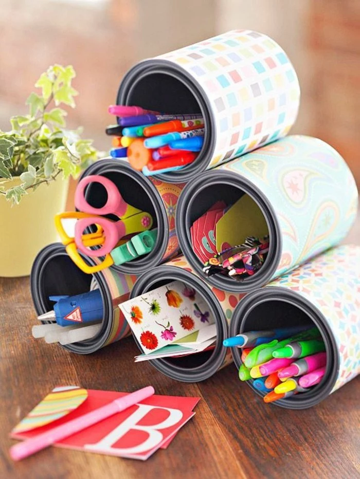 tin cans, six cans stacked in pyramid shape, decorated with multicolored patterned paper, containing pens and pencils, stickers and various stationary