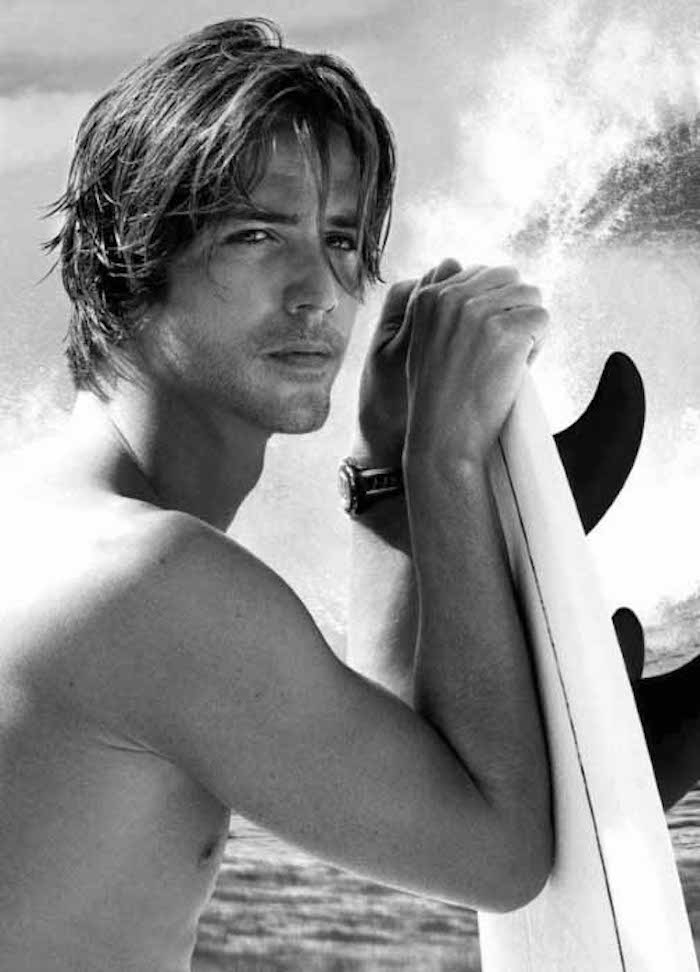 topless man leaning on surfboard, with wet layered hair, strands falling on his face, sea waves in the background