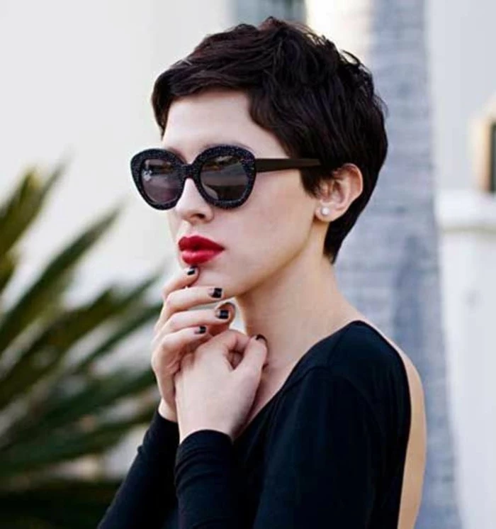 woman with very short dark wavy hair, wearing black retro sunglasses, bright red lipstick, and black open back top