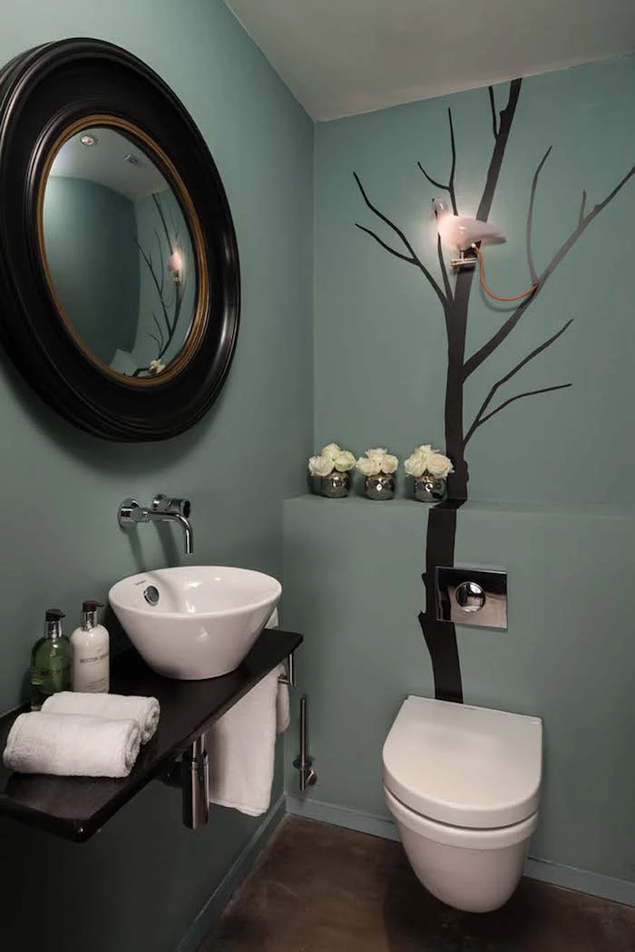bathroom decorating ideas, toilet with walls in duck egg's blue, one wall is decorated by a mural of black leafless tree, round mirror with large dark wooden frame hangs on other visible wall