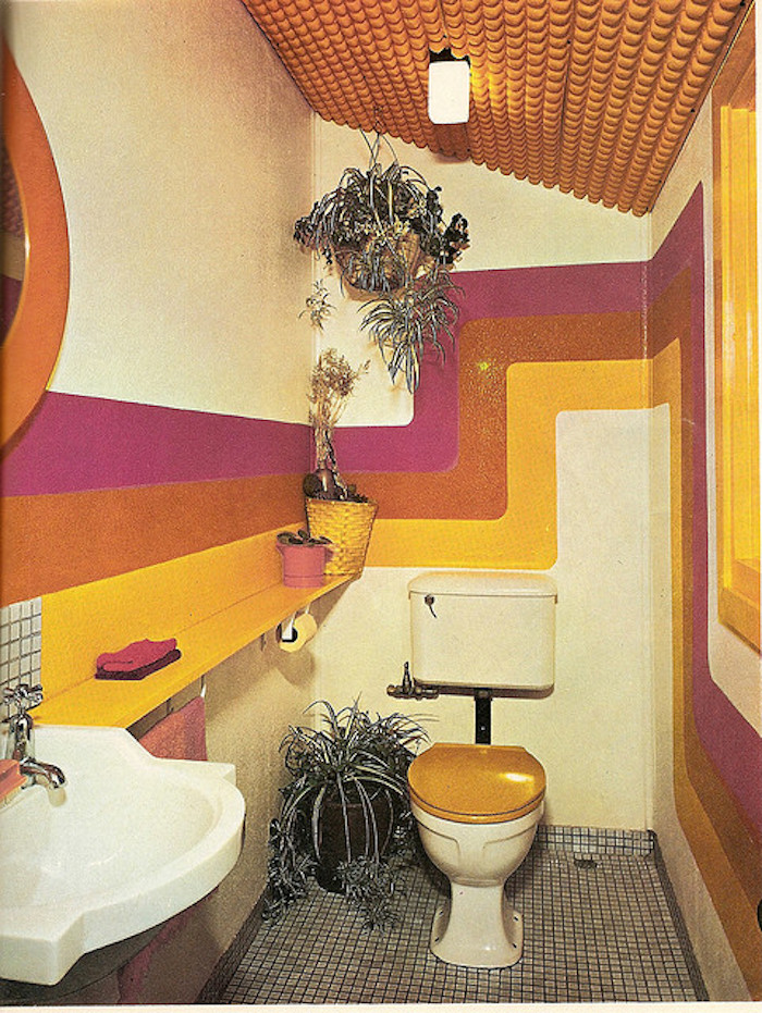 retro 1960's toilet, grey mosaic floor, white retro sink, toilet seat with wooden cover, walls with pink, orange and yellow bending stripes, several potted plants, orange textured ceiling