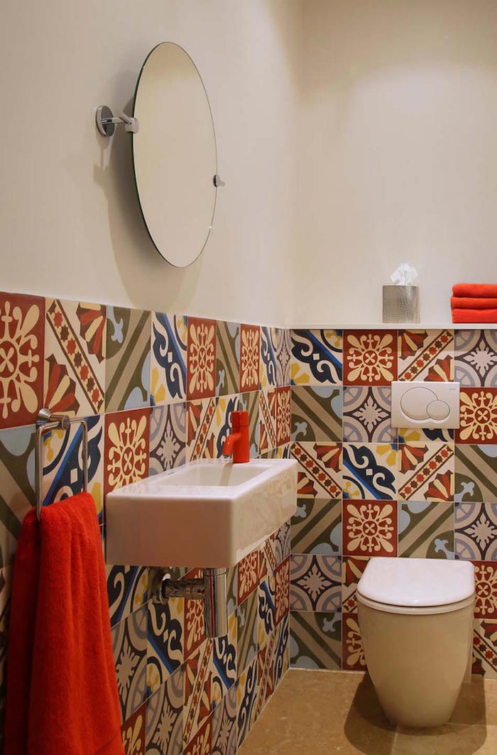 bathroom decorating ideas, bathroom with modern white toilet seat, and rectangular sink, red water tap and towels, upper half of the walls is plain white, lower part is decorated by multicolored, patterned and mismatched tiles