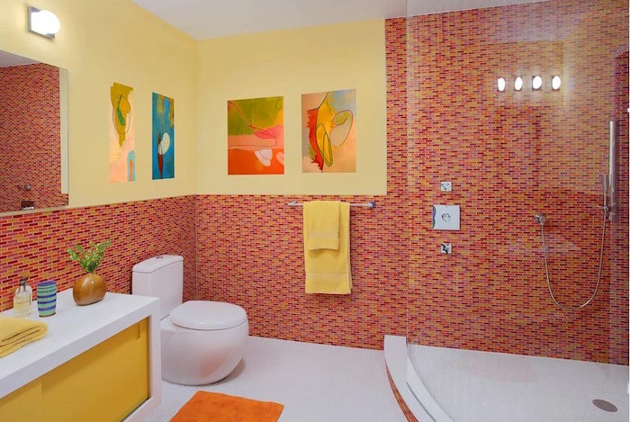 bathroom ideas, large room with big glass shower cabin, white and yellow slide cupboard, round modern white toilet seat, walls half covered with red, orange and yellow mosaic, other half painted in yellow, large mirror and paintings on walls